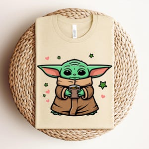 May include: Beige t-shirt featuring a cartoon illustration of a green alien character holding a cup. The character is wearing a brown cloak and has large ears. Pink hearts and green stars surround the character.