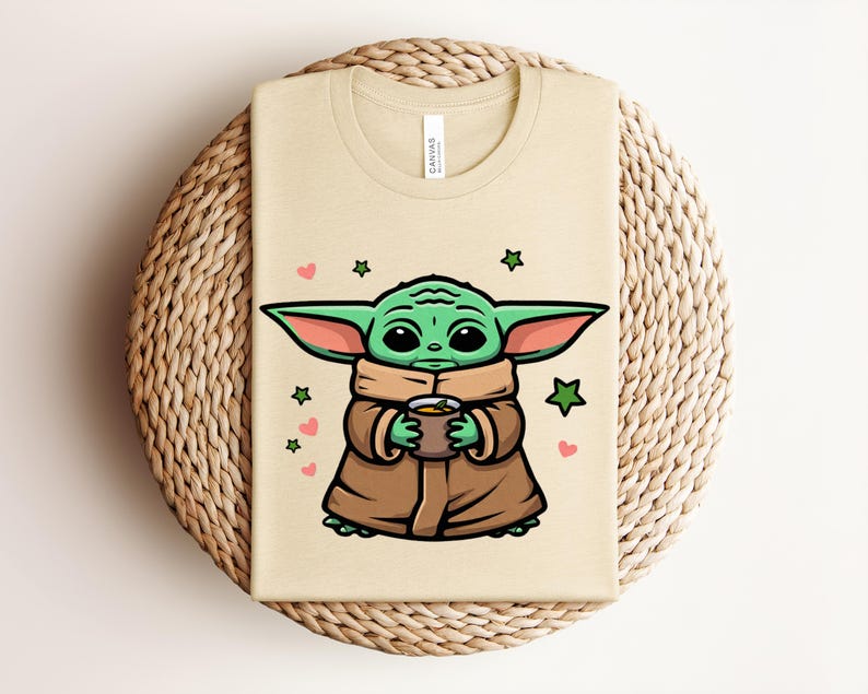 May include: Beige t-shirt featuring a cartoon illustration of a green alien character holding a cup. The character is wearing a brown cloak and has large ears. Pink hearts and green stars surround the character.