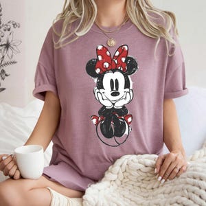 May include: Mauve t-shirt with a black and white Minnie Mouse illustration. Minnie is wearing a red polka-dot bow. The shirt has rolled-up sleeves and a relaxed fit. The image is of a cartoon character.