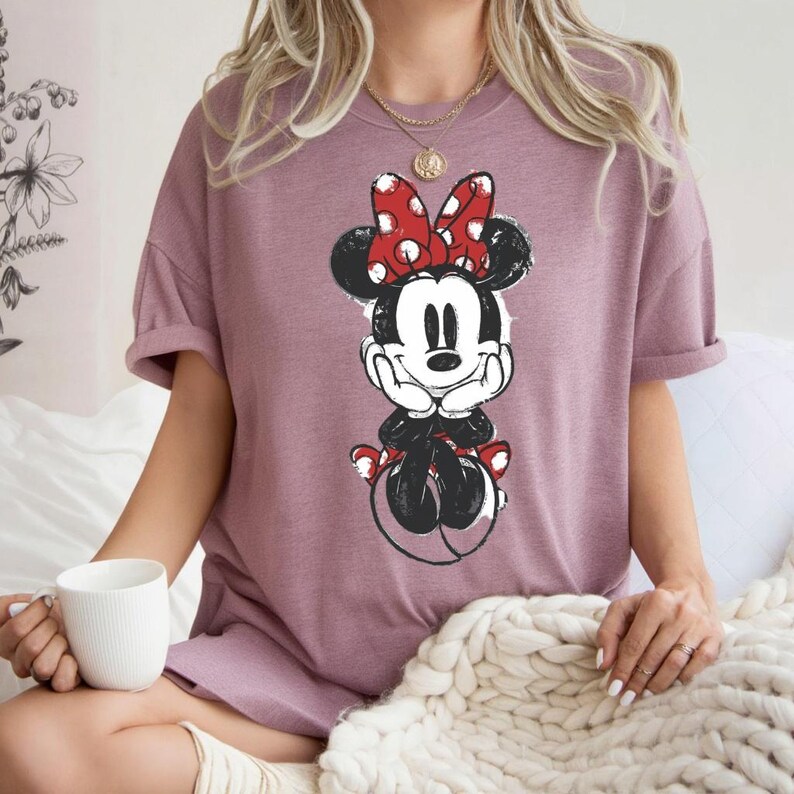 May include: Mauve t-shirt with a black and white Minnie Mouse illustration. Minnie is wearing a red polka-dot bow. The shirt has rolled-up sleeves and a relaxed fit. The image is of a cartoon character.