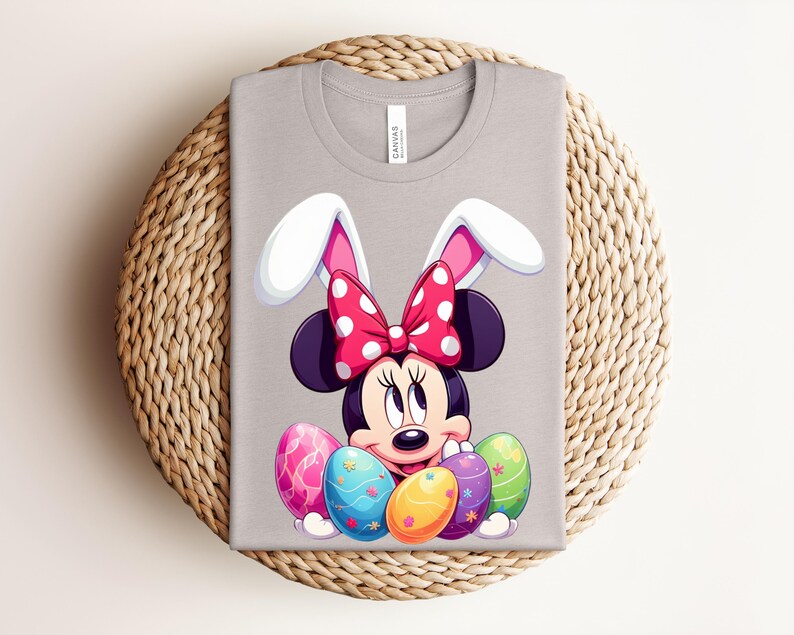 May include: A light grey t-shirt featuring a cartoon Minnie Mouse character with bunny ears and a red polka dot bow. She is surrounded by colourful Easter eggs. The shirt is folded on a woven surface.