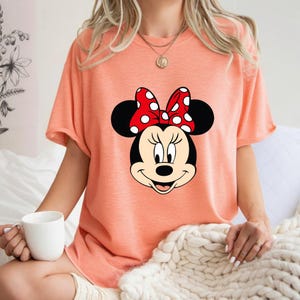 Op de afbeelding: Perzikkleurig T-shirt met een grote afbeelding van het gezicht van Minnie Mouse. De afbeelding bevat zwarte oren, een rode strik met witte stippen en een lachend gezicht. Het shirt heeft korte mouwen.