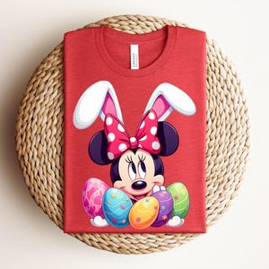 May include: Red t-shirt featuring Minnie Mouse dressed as an Easter bunny, with white bunny ears and a polka-dot bow. Minnie is surrounded by colourful Easter eggs. The shirt is folded on a woven, circular placemat.