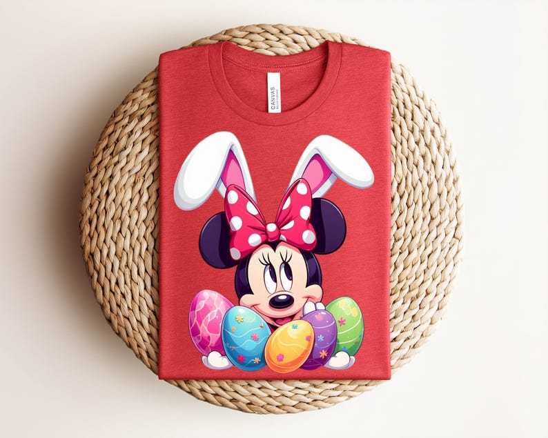 May include: Red t-shirt featuring Minnie Mouse dressed as an Easter bunny, with white bunny ears and a polka-dot bow. Minnie is surrounded by colourful Easter eggs. The shirt is folded on a woven, circular placemat.