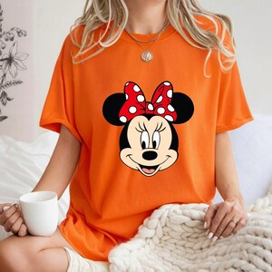 Op de afbeelding: Oranje T-shirt met een Minnie Mouse-afbeelding. De afbeelding toont Minnie's gezicht met een rode strik met witte stippen. Het shirt heeft korte mouwen en een relaxte pasvorm.