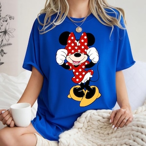 K&ouml;nnte beinhalten: Ein k&ouml;nigsblaues T-Shirt mit einem Minnie Mouse-Motiv. Die Zeichentrickfigur tr&auml;gt ihr klassisches Outfit: ein rotes Kleid mit wei&szlig;en Punkten und eine Schleife, wei&szlig;e Handschuhe und gelbe Schuhe. Minnie Mouse l&auml;chelt mit erhobenen H&auml;nden in der N&auml;he ihres Gesichts.