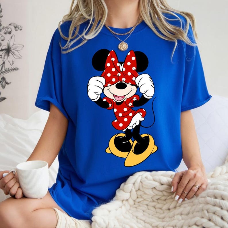 K&ouml;nnte beinhalten: Ein k&ouml;nigsblaues T-Shirt mit einem Minnie Mouse-Motiv. Die Zeichentrickfigur tr&auml;gt ihr klassisches Outfit: ein rotes Kleid mit wei&szlig;en Punkten und eine Schleife, wei&szlig;e Handschuhe und gelbe Schuhe. Minnie Mouse l&auml;chelt mit erhobenen H&auml;nden in der N&auml;he ihres Gesichts.