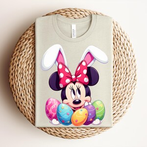 May include: A light beige t-shirt featuring a cartoon Minnie Mouse character wearing bunny ears and a red polka dot bow. The design includes colourful Easter eggs. The shirt is folded on a woven, circular surface.