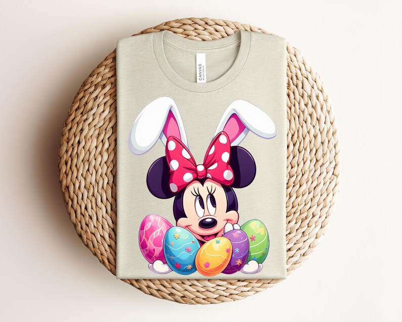 May include: A light beige t-shirt featuring a cartoon Minnie Mouse character wearing bunny ears and a red polka dot bow. The design includes colourful Easter eggs. The shirt is folded on a woven, circular surface.
