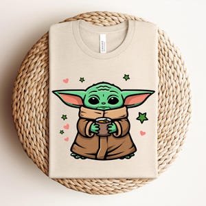 May include: Beige t-shirt featuring a cartoon illustration of a green alien character holding a cup. The design includes green stars and pink hearts. The shirt is folded on a woven, circular surface.