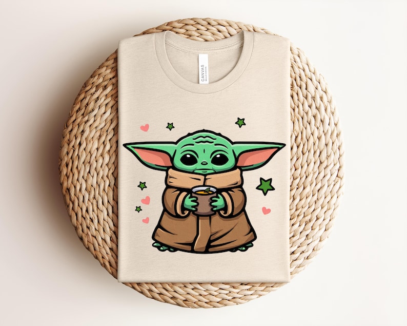 May include: Beige t-shirt featuring a cartoon illustration of a green alien character holding a cup. The design includes green stars and pink hearts. The shirt is folded on a woven, circular surface.