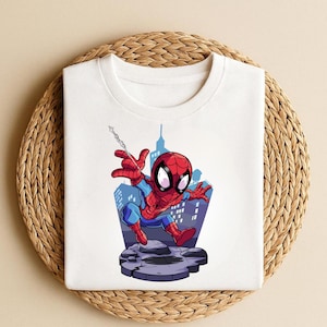 K&ouml;nnte beinhalten: Wei&szlig;es T-Shirt mit einem Cartoon-Design von Spider-Man. Die Illustration zeigt den Superhelden in seinem rot-blauen Kost&uuml;m, mit einem Stadtlandschaftshintergrund. Die Grafik ist mittig auf dem T-Shirt platziert.