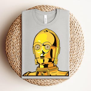 May include: A light gray t-shirt featuring a graphic of C-3PO from Star Wars. The illustration depicts the iconic golden droid's head and upper torso. The design is rendered in shades of yellow and black, with detailed linework.