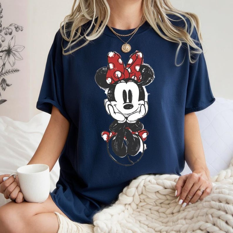 May include: Navy blue t-shirt with a graphic of Minnie Mouse. The cartoon character is in black and white with a red polka dot bow. The shirt has a relaxed fit and short sleeves.