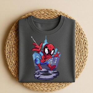K&ouml;nnte beinhalten: Dunkelgraues T-Shirt mit einem Spider-Man-Cartoon-Motiv. Der Superheld ist in seinem klassischen rot-blauen Anzug vor einer Stadtkulisse dargestellt. Das Design ist auf einem Rundhals-T-Shirt gedruckt, ideal f&uuml;r Freizeitkleidung.