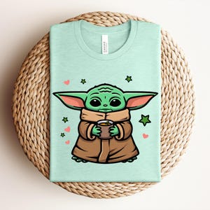 May include: A light green t-shirt featuring a cartoon character with large ears, holding a cup. The character is green with brown clothing, surrounded by hearts and stars. The shirt is folded on a woven surface.
