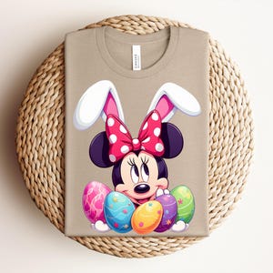 May include: Tan t-shirt featuring a cartoon Minnie Mouse design with bunny ears and a red polka dot bow. The design includes colourful Easter eggs. The shirt is folded on a woven mat.