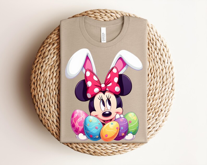 May include: Tan t-shirt featuring a cartoon Minnie Mouse design with bunny ears and a red polka dot bow. The design includes colourful Easter eggs. The shirt is folded on a woven mat.
