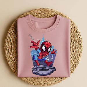 K&ouml;nnte beinhalten: Ein hellrosa Sweatshirt mit Rundhalsausschnitt und einer Cartoon-Illustration von Spider-Man. Der Superheld ist in seinem klassischen rot-blauen Anzug zu sehen, wie er vor einer Stadtkulisse an einem Netz schwingt. Das Sweatshirt liegt auf einer gewebten Matte.