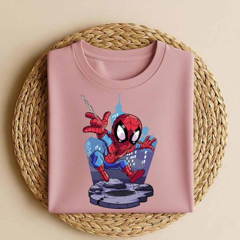 K&ouml;nnte beinhalten: Ein hellrosa Sweatshirt mit Rundhalsausschnitt und einer Cartoon-Illustration von Spider-Man. Der Superheld ist in seinem klassischen rot-blauen Anzug zu sehen, wie er vor einer Stadtkulisse an einem Netz schwingt. Das Sweatshirt liegt auf einer gewebten Matte.