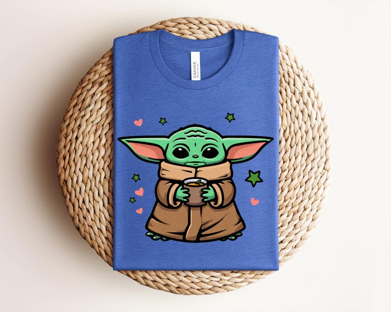 May include: A blue t-shirt featuring a cartoon character with large green ears, holding a cup. The character is wearing a brown coat and is surrounded by hearts and stars. The shirt is folded on a woven surface.