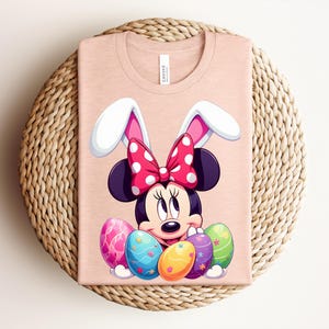 May include: A pale pink t-shirt featuring a cartoon Minnie Mouse design with bunny ears and a red polka dot bow. Colourful Easter eggs are at the bottom. The shirt is folded on a woven surface.