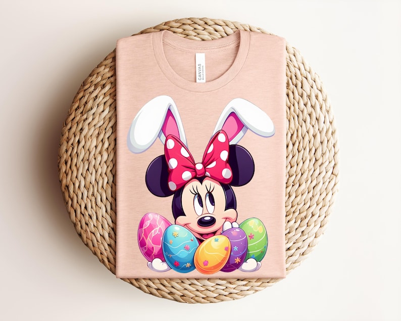 May include: A pale pink t-shirt featuring a cartoon Minnie Mouse design with bunny ears and a red polka dot bow. Colourful Easter eggs are at the bottom. The shirt is folded on a woven surface.