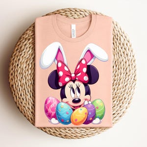 May include: A peach-coloured t-shirt featuring a cartoon Minnie Mouse design with bunny ears and a polka-dot bow. Minnie is surrounded by colourful Easter eggs. The shirt is folded on a woven, circular placemat.