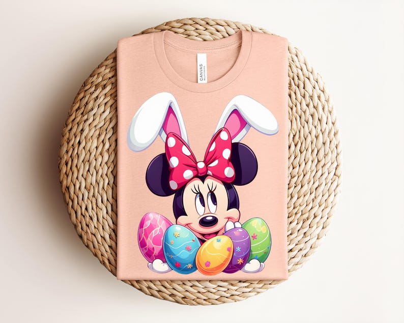 May include: A peach-coloured t-shirt featuring a cartoon Minnie Mouse design with bunny ears and a polka-dot bow. Minnie is surrounded by colourful Easter eggs. The shirt is folded on a woven, circular placemat.