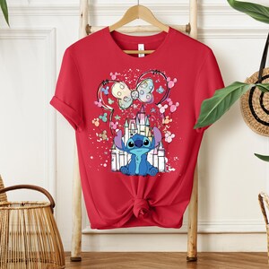 K&ouml;nnte beinhalten: Rotes T-Shirt mit einem farbenfrohen Aufdruck von Stitch, einem Schloss und einer Minnie-Mouse-Schleife. Das Design umfasst Pastellfarben und einen Spritzeffekt. Das T-Shirt ist an der Taille geknotet.