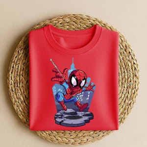 K&ouml;nnte beinhalten: Ein rotes Rundhals-T-Shirt mit einer Cartoon-Illustration von Spider-Man. Der Superheld ist in seinem rot-blauen Anzug dargestellt, mit einer Stadtkulisse. Das T-Shirt ist auf einer gewebten Matte gefaltet.