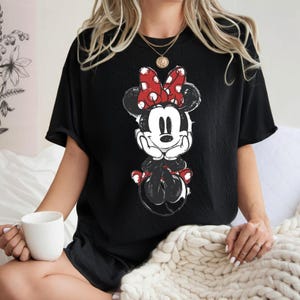 May include: Black t-shirt with a distressed Minnie Mouse graphic. The cartoon character is in black, white, and red, with a large polka-dot bow. The shirt has short sleeves and a relaxed fit, suitable for casual wear.
