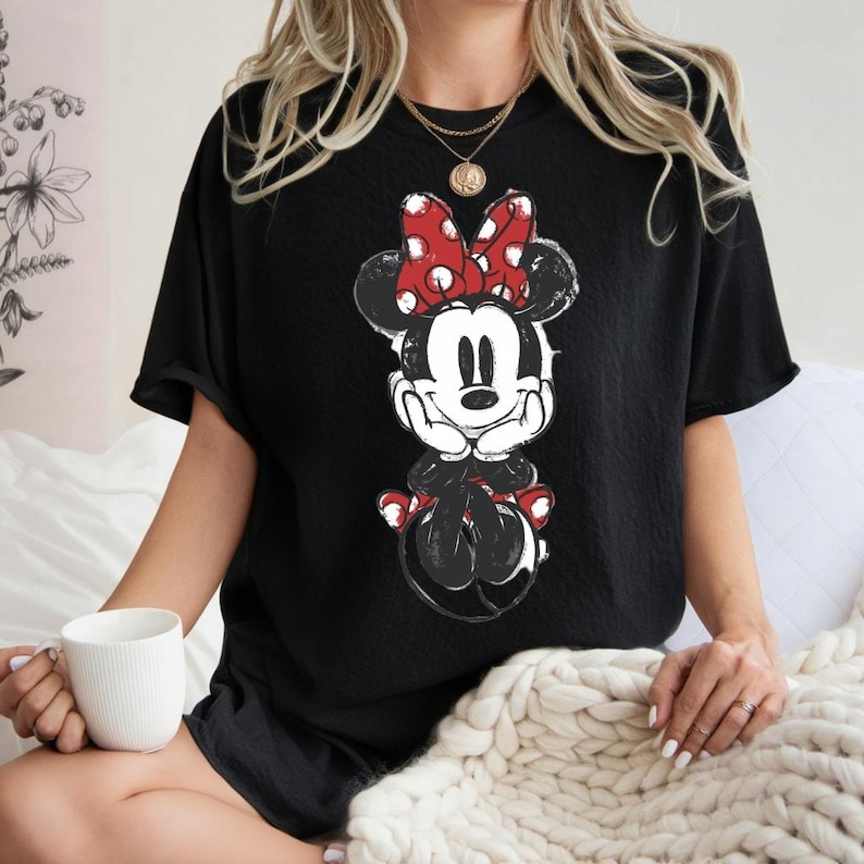 May include: Black t-shirt with a distressed Minnie Mouse graphic. The cartoon character is in black, white, and red, with a large polka-dot bow. The shirt has short sleeves and a relaxed fit, suitable for casual wear.