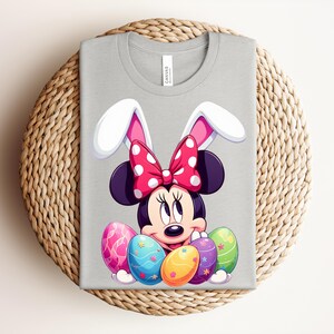 May include: A light grey t-shirt featuring a cartoon Minnie Mouse character with bunny ears and a red polka dot bow. The design includes colourful Easter eggs. The shirt is displayed on a woven, circular surface.