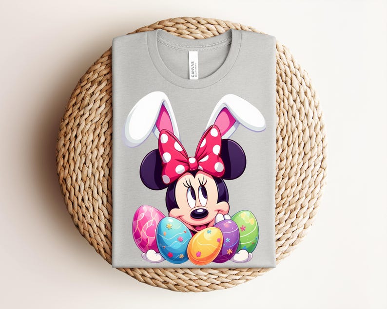 May include: A light grey t-shirt featuring a cartoon Minnie Mouse character with bunny ears and a red polka dot bow. The design includes colourful Easter eggs. The shirt is displayed on a woven, circular surface.