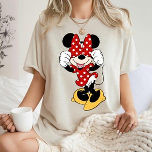 K&ouml;nnte beinhalten: Beiges T-Shirt mit Minnie Mouse-Grafik. Die Zeichentrickfigur tr&auml;gt ein rotes Kleid mit wei&szlig;en Punkten, eine passende Schleife und gelbe Schuhe. Das Design ist mittig auf der Vorderseite des Shirts platziert.