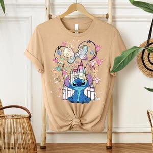 K&ouml;nnte beinhalten: Beiges T-Shirt mit einem Cartoon-Stitch-Design vor einem Schloss. Das Design umfasst eine Mickey-Mouse-Schleife und farbenfrohe Akzente. Das T-Shirt h&auml;ngt an einem Holzb&uuml;gel.