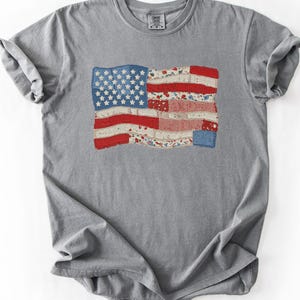 May include: A gray t-shirt featuring a distressed American flag design. The flag has a patchwork style with red and white stripes and a blue field of stars. The shirt is made of a soft material.