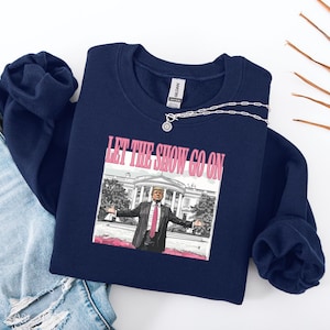 May include: Navy blue sweatshirt with the text "LET THE SHOW GO ON" in pink and a graphic of a man in a suit with the White House in the background. A silver necklace with a pendant is draped across the sweatshirt.