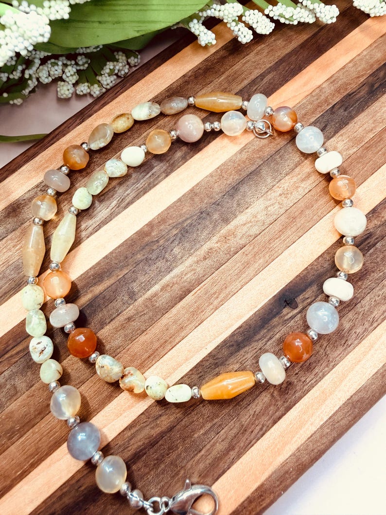 May include: A beaded necklace featuring a variety of smooth, polished stones in shades of orange, cream, pale yellow, and translucent gray. The beads are interspersed with small, round silver-toned metal spacers and secured with a silver-toned lobster clasp.
