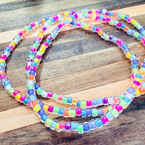 May include: A multi-strand necklace composed of small, translucent, cylindrical beads in a vibrant array of colours including pink, yellow, orange, green, blue, and purple. The beads are strung together in a repeating pattern, creating a bright, colourful accessory that loops in several circular layers.
