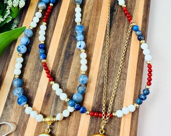 May include: Two layered necklaces featuring blue, white, and red beads with gold-tone accents. One necklace has a pendant with a blue sailboat illustration, and the other features a pendant with a blue seashell design on a white background.