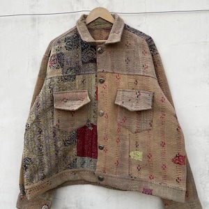 Op de afbeelding: Een jas met lange mouwen en knoopsluiting, gemaakt van patchworkstof met een mix van beige, bruine en veelkleurige patronen. Het kledingstuk is voorzien van twee borstzakken met kleppen, een standaard kraag en zichtbare stiksels in kantha-stijl.