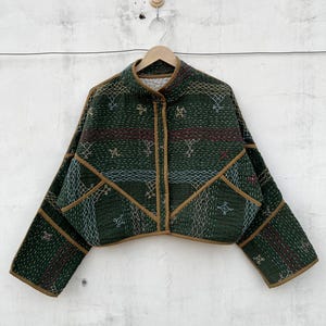 Puede incluir: Una chaqueta acolchada de color verde oscuro con cuello alto, mangas largas y ribetes de color tostado en los bordes. La tela está decorada con intrincados patrones geométricos bordados en blanco, amarillo y rojo y costuras kantha.