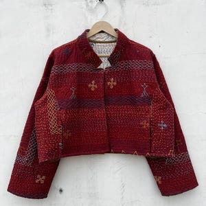 May include: A cropped jacket in deep red featuring intricate white and multicolored kantha embroidery. The garment has long sleeves, a stand-up collar, and a quilted texture with visible running stitches throughout the fabric surface.