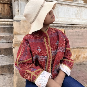 Op de afbeelding: Een beige bucket hat met een dunne band en een langarmig, gewatteerd jack in donkerrood met ingewikkeld kantha-borduurwerk in blauwe, witte en gele patronen, gecombineerd met een donkerblauwe spijkerbroek, met een mandarijnkraag en manchetten.