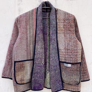 Könnte beinhalten: Mehrfarbige Jacke mit offenem Schnitt und geometrischem Muster. Sie zeigt Lila-, Rosa- und Beigetöne mit dunkelblauen Paspeln. Zwei Taschen sind vorhanden. Auf einem Metallbügel an einer strukturierten weißen Wand.