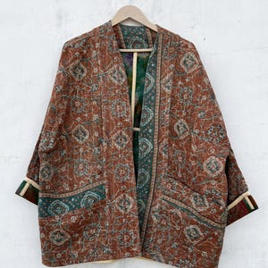 Puede incluir: Una chaqueta acolchada con un patrón floral geométrico en color óxido y verde azulado con costuras kantha visibles. La prenda abierta por delante tiene mangas tres cuartos con puños en contraste y dos bolsillos de parche en la parte frontal.