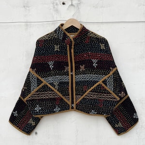 May include: A black quilted jacket featuring a high collar and tan piping along the edges. The fabric is decorated with horizontal bands of intricate white, red, and tan geometric embroidery and stitching patterns across the front and sleeves.