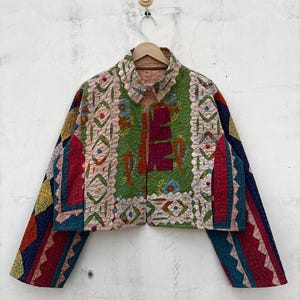 Pode incluir: Um casaco curto com design de patchwork e texturas acolchoadas. A peça exibe uma mistura vibrante de painéis verdes, rosa, azuis e vermelhos com intrincados padrões de bordado geométricos e florais em toda a superfície do tecido.